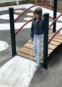 Lapsi kävelee valkoisen kepin kanssa leikkipuiston sillalla, asfaltissa valkoista maalia.