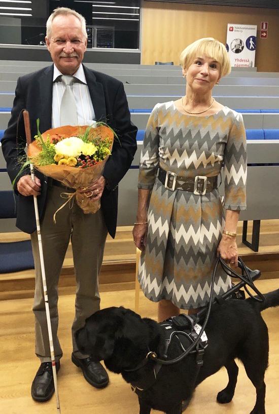 Mies valkoisen kepin ja kukkakimpun kanssa ja nainen, jolla musta opaskoira. Molemmat hymyilevät.