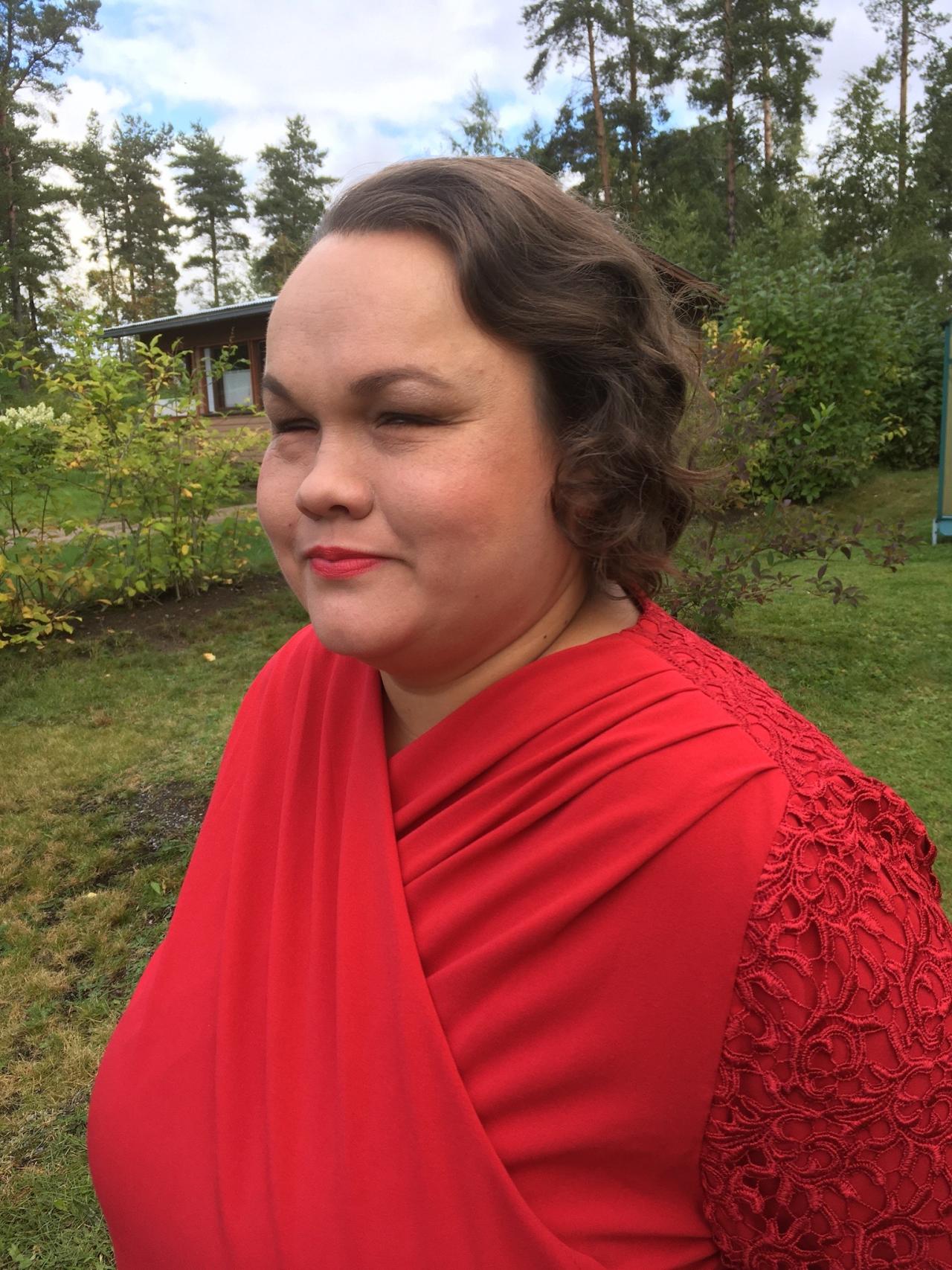 Sivukuva Päivistä rinnasta ylöspäin punaisessa mekossa pihalla. Taustalla talo ja vihreä kesäpuutarha. 