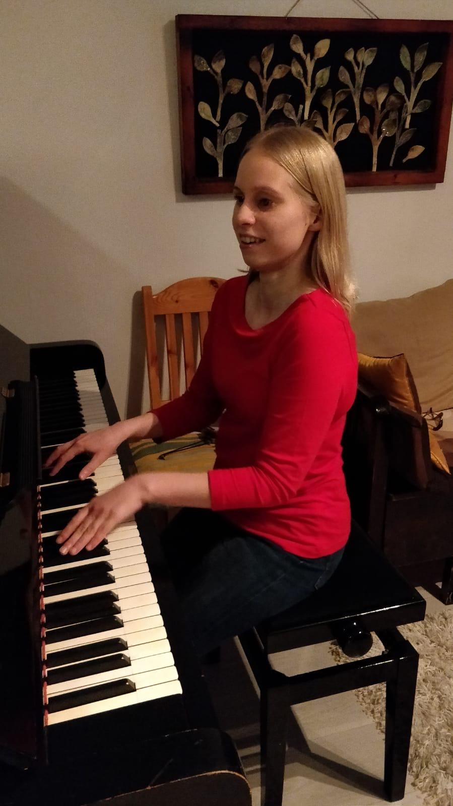 Minja soittaa mustaa pianoa ja hymyilee. Minjalla on punainen paita ja taustalla näkyy puinen tuoli ja sohvaa.