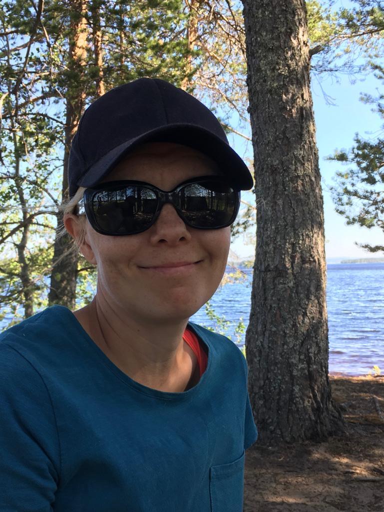 Maija katsoo hymyillen kameraan kesäisessä järvimaisemassa. Päässään Maijalla on aurinkolasit ja lippalakki.