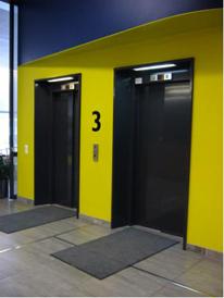 Keltainen seinä, jonka keskellä kaksi mustaovista hissiä ja iso numero 3. Molemmille hissinoville johtaa tumma lista.