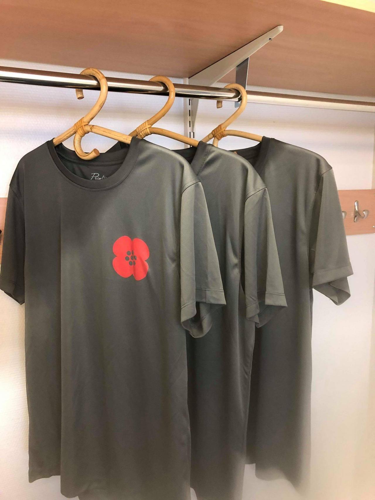 3 harmaata t-paitaa henkareissa naulakossa, paidan rinnassa punainen annansilmä-kukka.