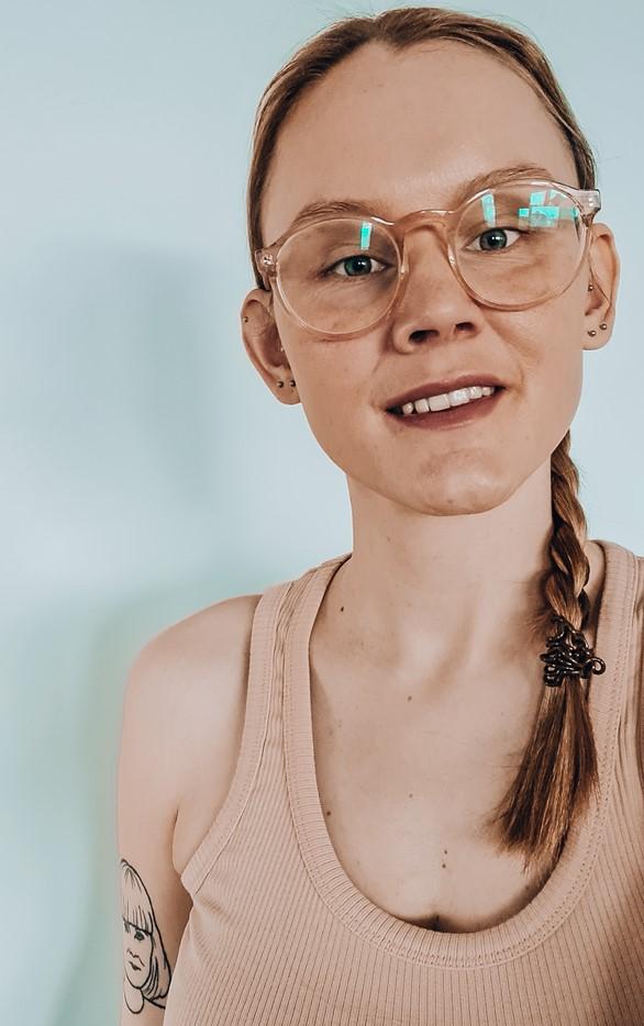 Hilma seisoo vaaleaa seinää vasten ja hymyilee. Hilmalla on hiukset letitetty ja silmälasit päässä.