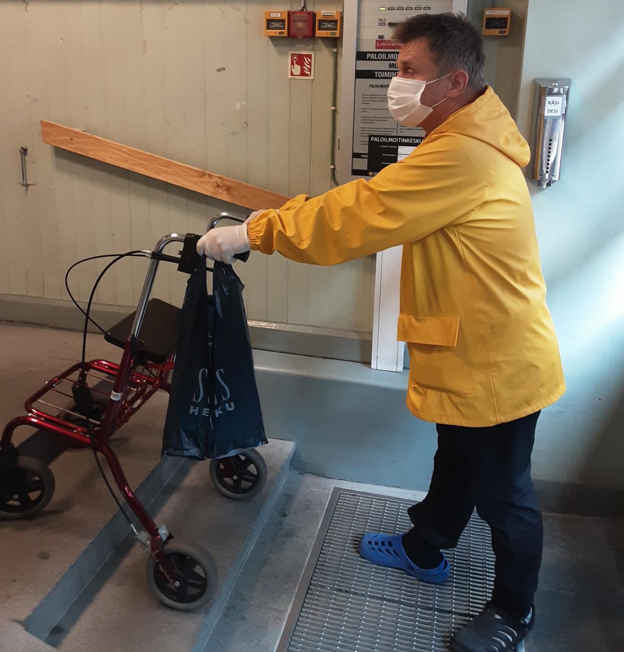 Markus kulkemassa rollaattorin kanssa porraskäytävässä. Portaita on kolme. Rollaattorin etupyörät on toisella portaalla. Markuksella on pirteän keltainen sadetakki ja maski. Rollaattorin kädensijassa roikkuu muovikassi.