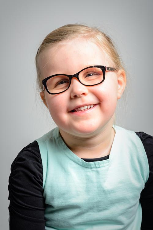 Muotokuva 4-vuotiaasta hymyilevästä silmälasipäisestä tytöstä. Tytöllä on vaaleat hiukset poninhännällä ja tummasankaiset silmälasit. Hänellä on vaalean turkoosin värinen tunika ja musta pitkähihainen aluspaita.