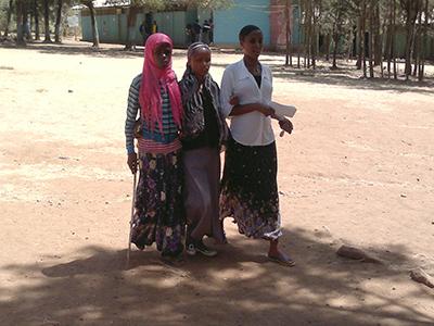 Kolme nuorta etiopialaisnaista kävelevät vierekkäin hiekkatiellä. Taustalla näkyy puita ja sininen talo.