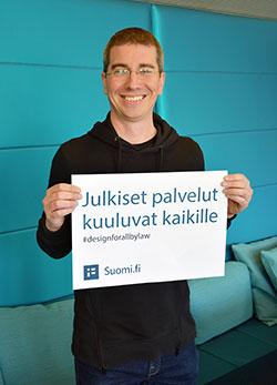 Jani Ruuskanen seisoo turkoosin seinän edessä, hänellä on silmälasit, lyhyt tumma tukka ja musta paita. Hän pitelee käsissään kylttiä, jossa lukee "Julkiset palvelut kuuluvat kaikille, #designforallbylaw, Suomi.fi".