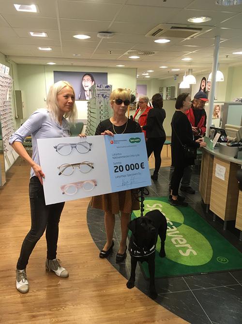 Vasemmalla Kaisa Mäkäräinen vaaleansinisessä paidassa ja oikealla Sari Loijas mustassa puserossa. He pitelevät välissään iso shekin näköistä julistetta, jossa näkyy summa 20 000 euroa.