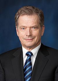 Tasavallan presidentti Sauli Niinistö, jolla on päällään tumma puku ja sininen solmio. Hänellä on vaaleanruskeat hiukset ja hän hymyilee.