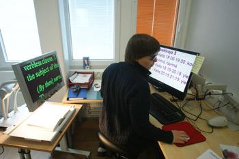 Nainen istuu kirjoituspöydän ääressä ja katsoo tietokoneen ruutua, jossa on suurennusohjelma. Vasemmalla on lukutelevisio.
