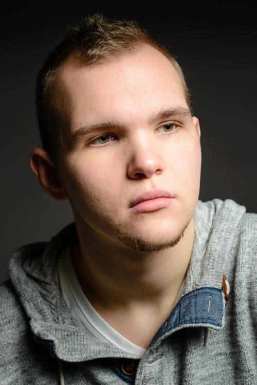 Muotokuva 22-vuotiaasta nuoresta miehestä. Hänellä onb vaaleanruskeat lyhyet hiukset ja vihertävänruskeat silmät. Hänellä on pieni leukaparta ja tummat kulmakarvat. Ilme on vakava. Yllään hänellä on harmaa neulehuppari.