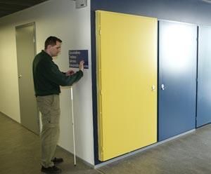 Käytävien risteyskohdassa on oikealla kirkkaankeltainen ovi ja vasemmalla mieshenkilö lukee pistekirjoitusta opastekyltissä.