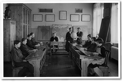 Mustavalkoinen kuva vanhasta koululuokasta. Keskellä on opettaja ja oppilaiden pulpetit ovat huoneen reunoilla.