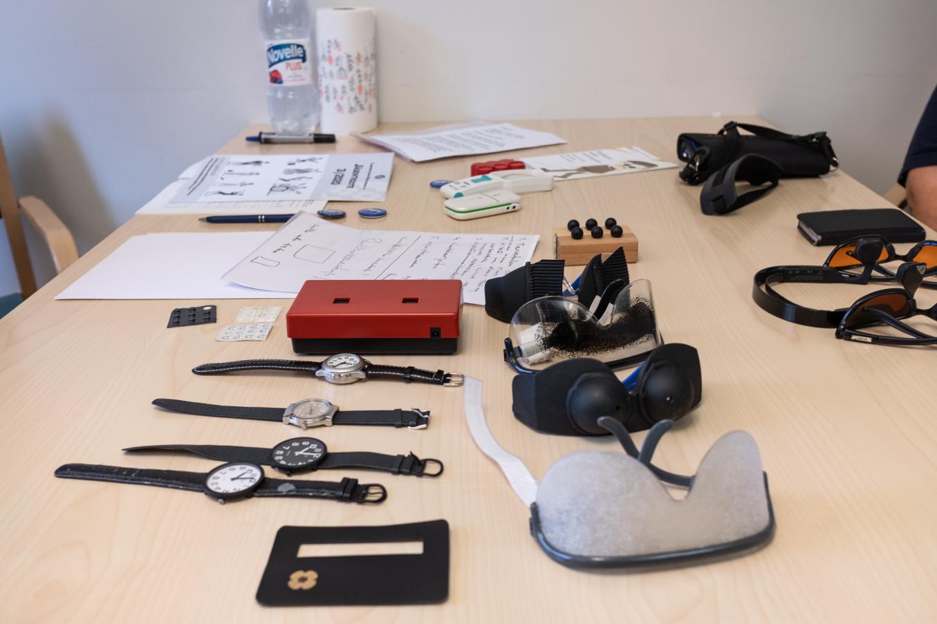 Pöydällä erilaisia rannekelloja, apuvälinelaseja ja muita tarvikkeita kokemuskoulutustilannetta varten.