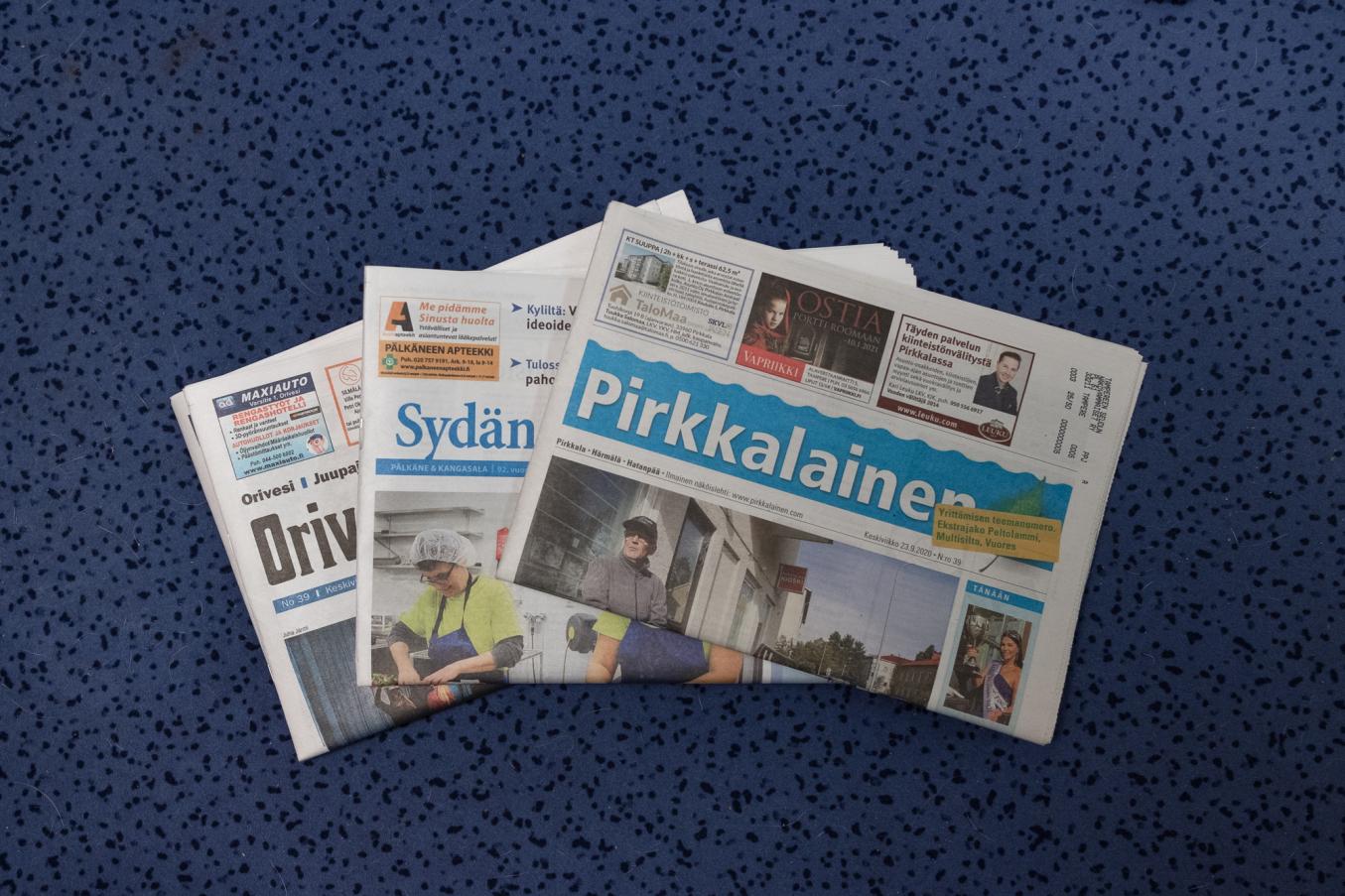 Sinisellä taustalla kolme paikallislehteä lomittain päällekkäin vinosti pinottuna. 