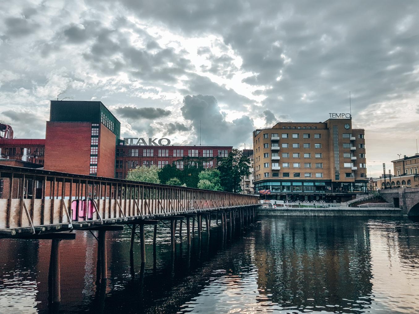 Kesäinen Takon tehdas ja Tempon talo kuvattuna toiselta puolelta rantaa. Kuvassa näkyy myös kosken ylittävä kävelysilta. 