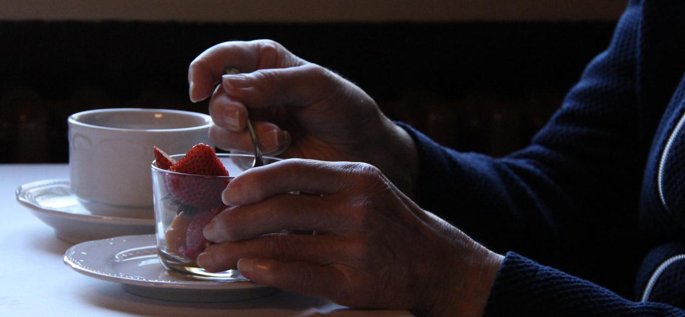 Hämärässä pöydällä kahvikuppi ja mansikoita lasikipossa. Nainen pitää kättään mansikkakipon ympärillä ja on ottamassa lusikalla mansikoita lasikiposta.