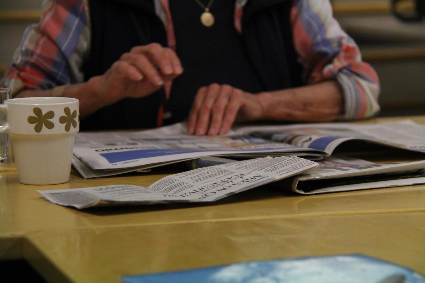 Nainen lukee sanomalehtiä pöydän ääressä, pöydällä lisäksi useita muita lehtiä ja kahvikuppi. 