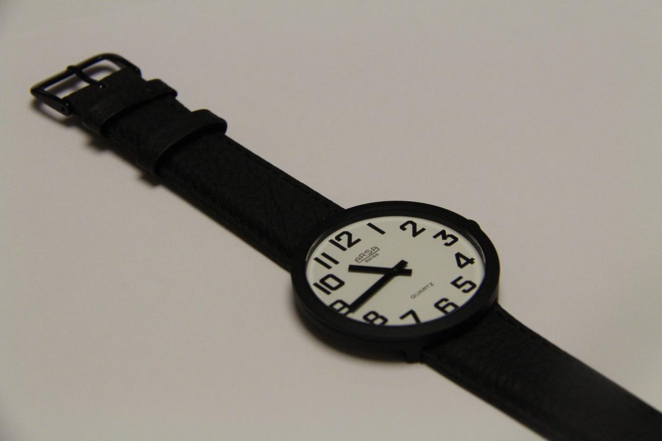 Valkoisella näytöllä ja mustilla viisareilla varustettu musta rannekello valkoisella pöydällä.