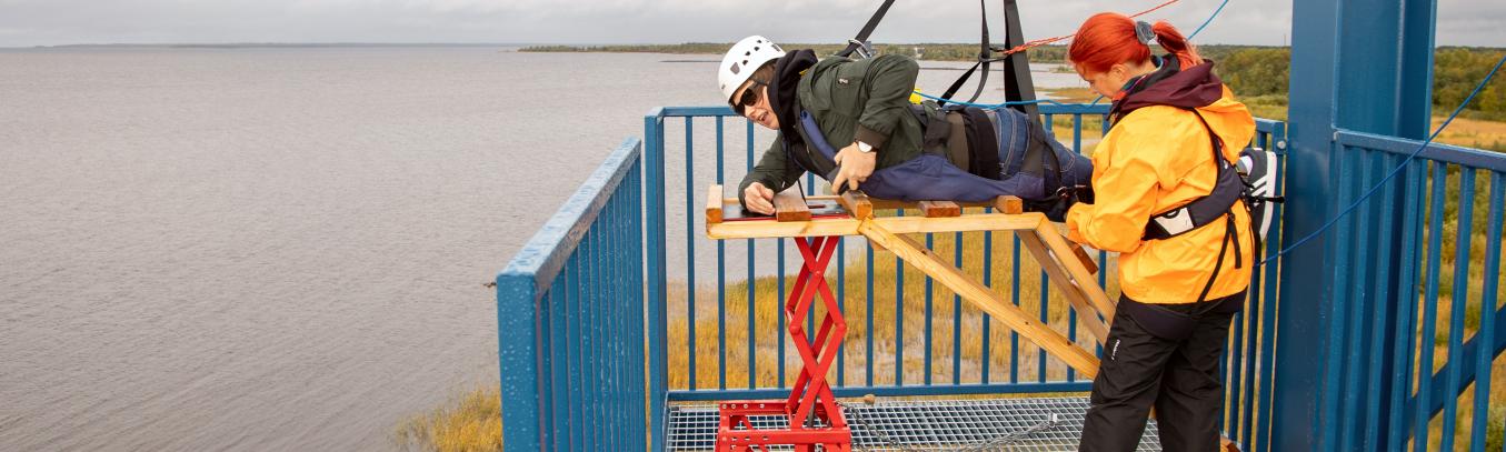 Retkeläinen valmistautumassa vaijeriliukuun ja järjestäjä laittaa turvallisuusremmejä paikoilleen