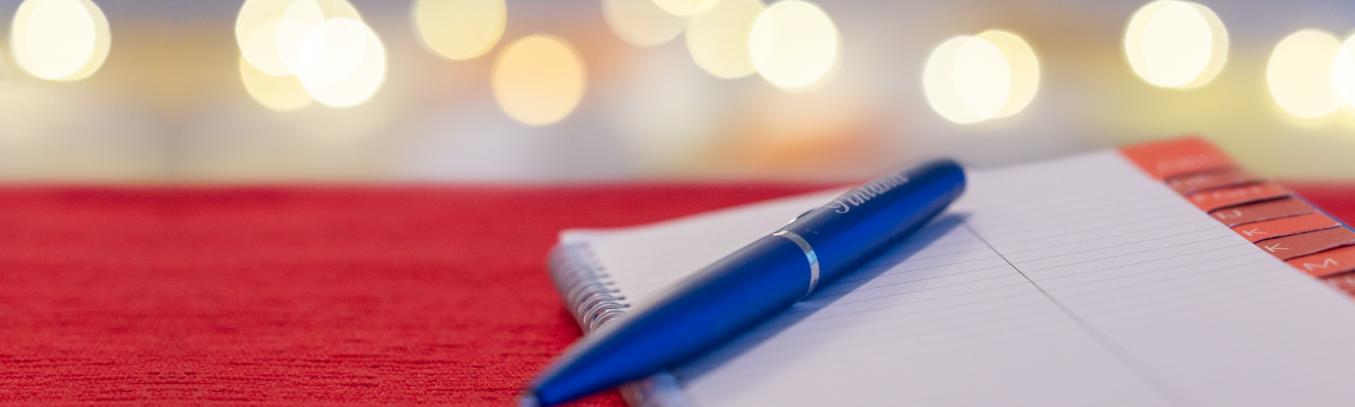 Kuvassa on osoitekirja ja kynä. Taustalla näkyy sumeita valopalloja