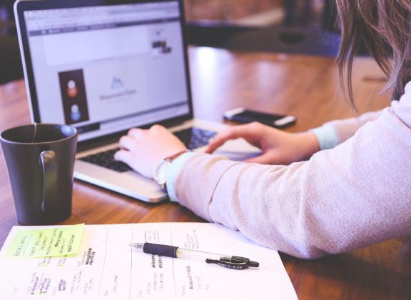 Nainen kirjoittaa tietokoneella. Pöydällä kahvikuppi, asiakirja, kynä ja puhelin.